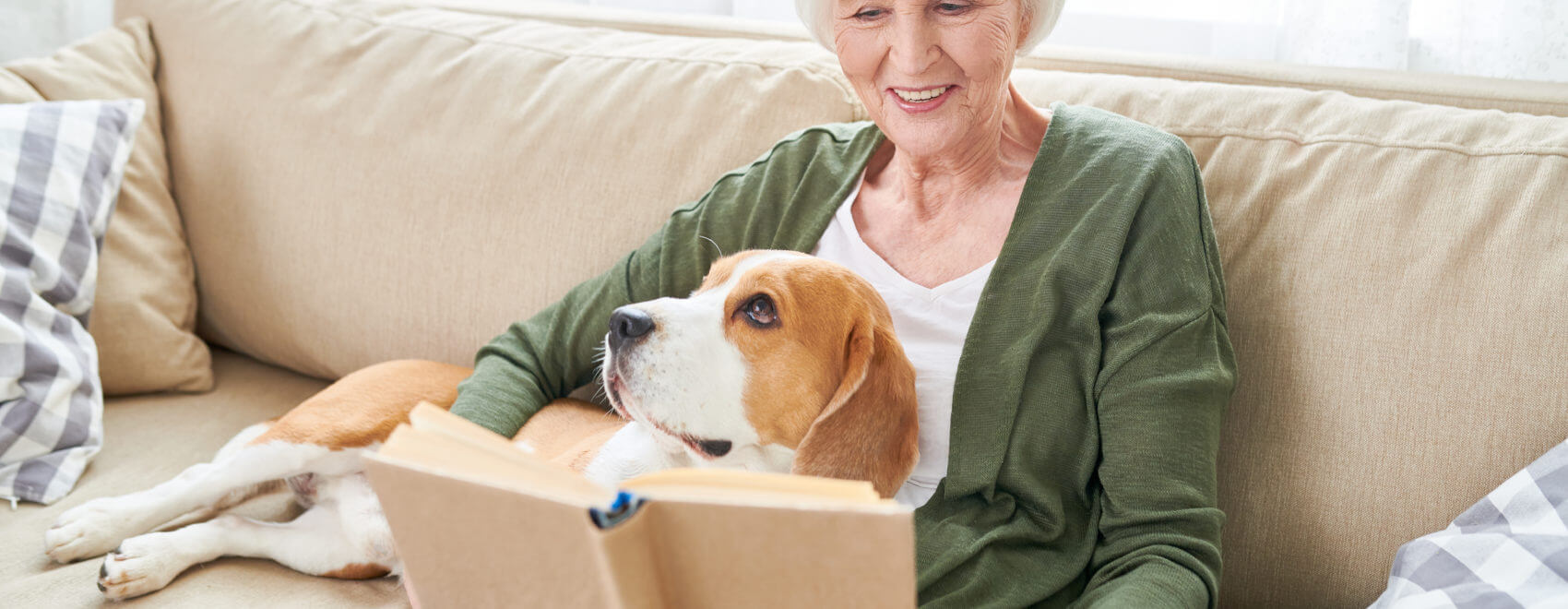 Portrait of happy senior woman enjoying leisure time at home sitting on comfortable couch with pet dog in her lap and reading books